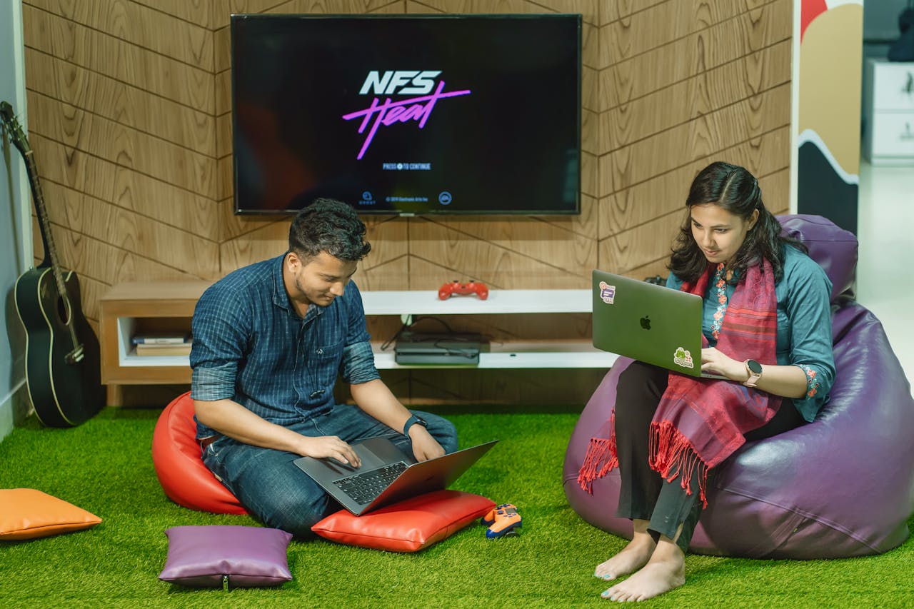 Two colleagues working on laptops in a relaxed office space with bean bags and a guitar.